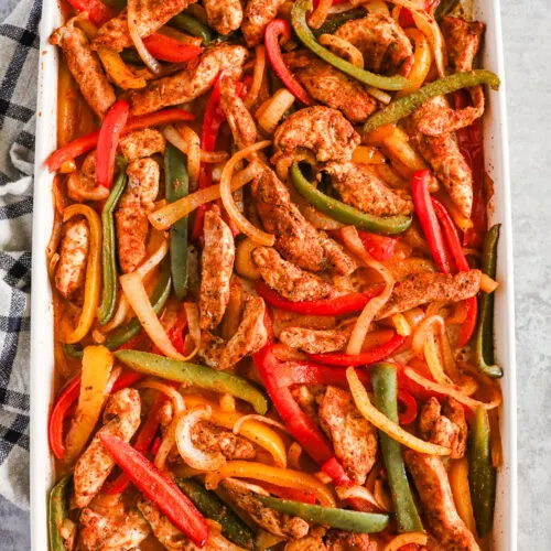 A baking sheet with thin strips of colorful bell pepper, onion and chicken, all covered in a taco seasoning.