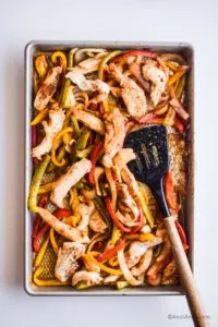 Sheet pan with chicken strips and sliced bell pepper and spatula.