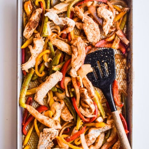 Sheet pan with chicken strips and sliced bell pepper and spatula.