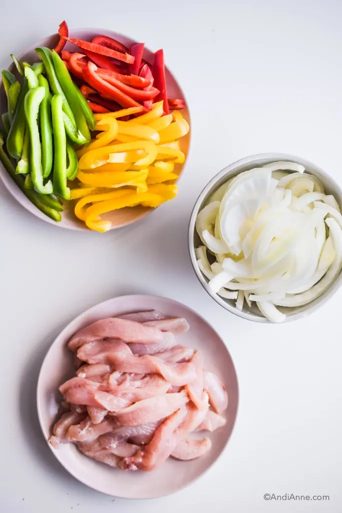 sliced red, green and yellow bell peppers on a plate. Sliced white onion in a grey bowl, and sliced raw chicken breast on a plate.