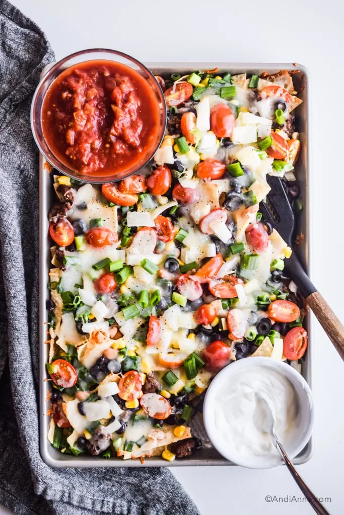 A baking sheet with nachos, tomato, olives, green onion, and melted cheese. A bowl of salsa and a bowl of sour cream. 
