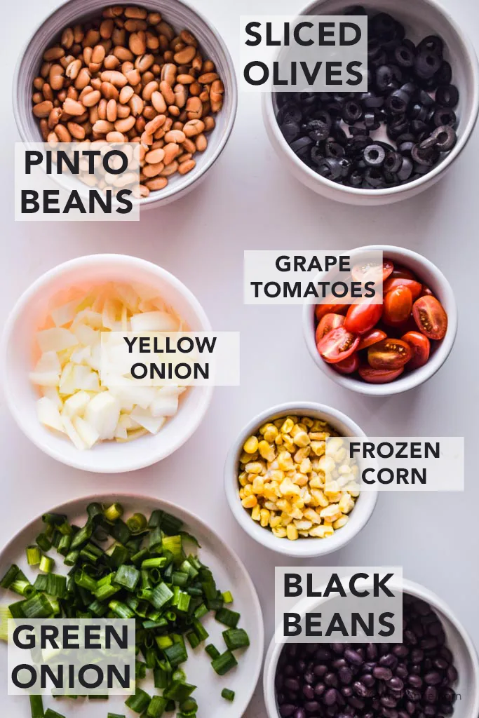 ingredients for sheet pan nachos in white bowls. Pinto beans, sliced olives, yellow onions, grape tomatoes, frozen corn, black beans and green onion