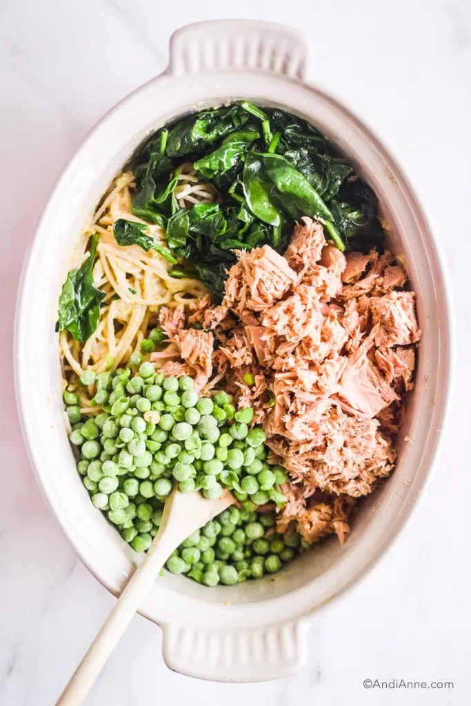 spinach, canned tuna, noodles, frozen peas inside a white bowl with a wooden spoon