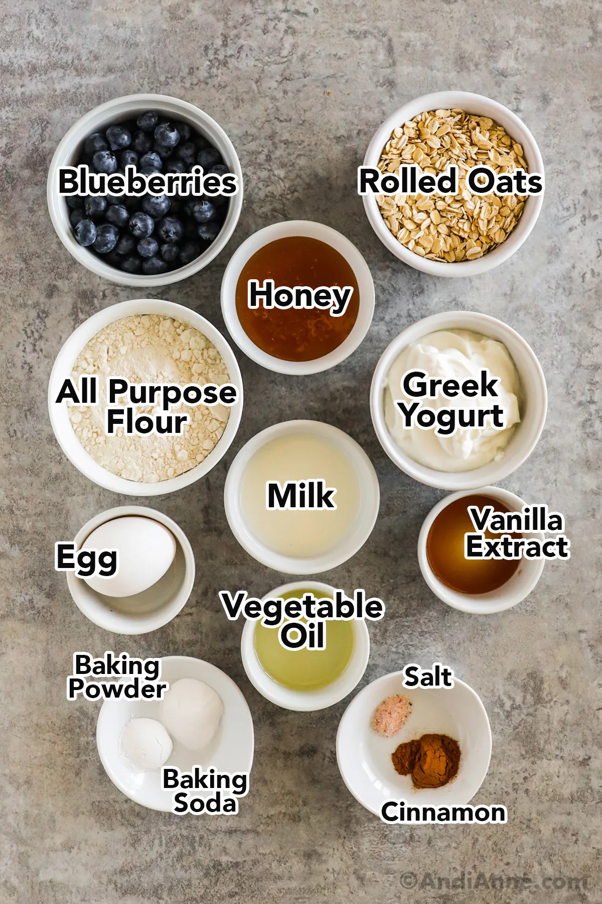 Recipe ingredients on the counter including bowls of blueberries, rolled oats, honey, flour, greek yogurt, milk, vanilla, vegetable oil, salt and baking powder.