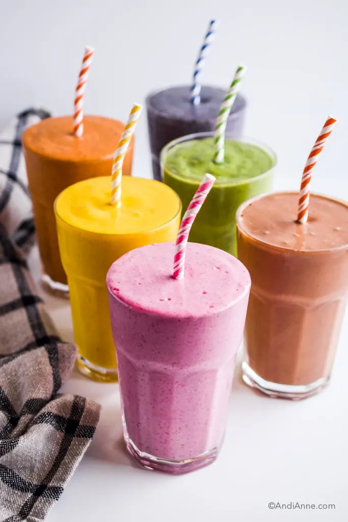 six healthy smoothies in glasses with paper straws: raspberry yogurt, banana chocolate, mango turmeric, green smoothie, blueberry, and carrot cake