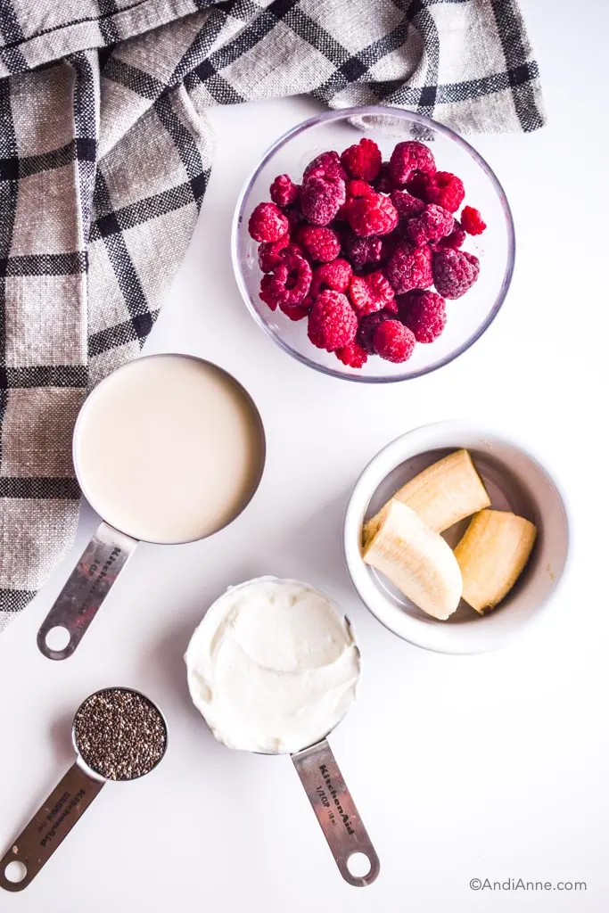 Glass bowl of raspberries, bowl of sliced frozen banana, measuring cups of milk, yogurt, and chia seeds. Kitchen napkin beside it all.