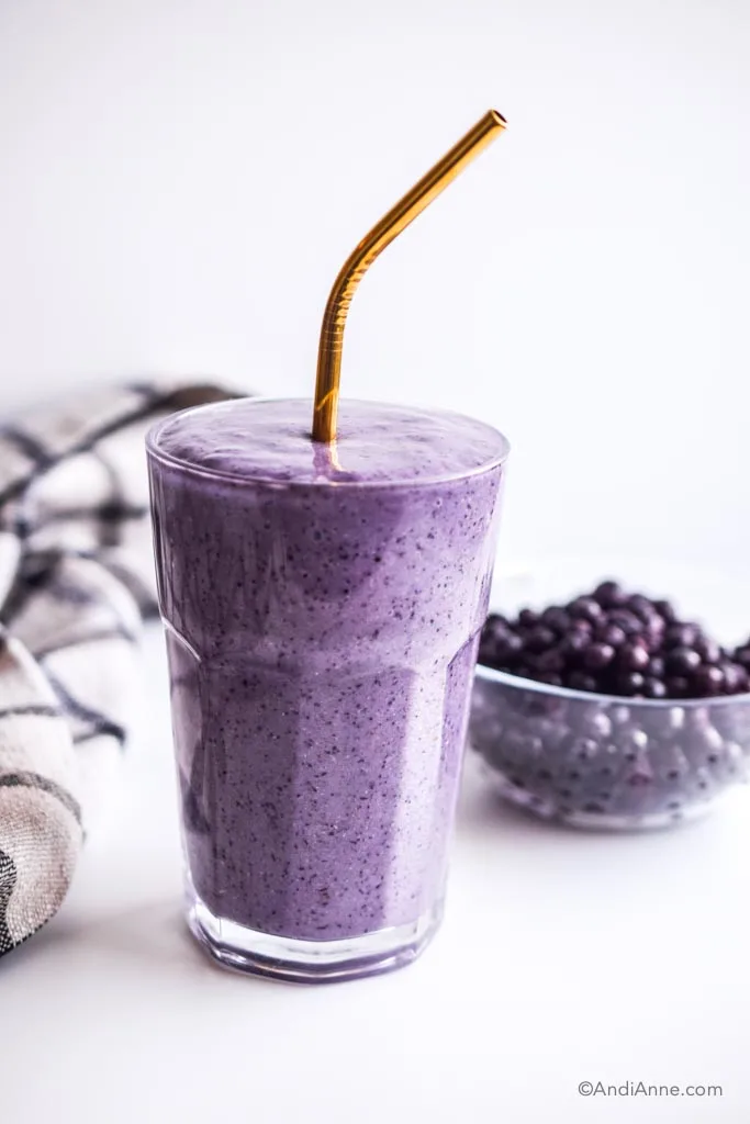 blueberry smoothie in a glass with metal straw and a bowl of blueberries behind it