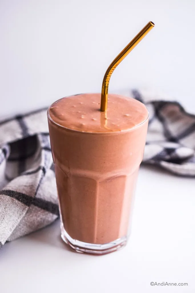 banana chocolate protein smoothie in a glass with gold straw. Kitchen napkin is behind it.