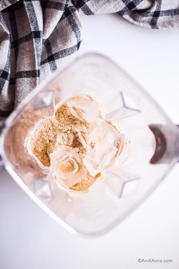 looking down into a blender cup wiht protein powder, ice cubes and other ingredients inside