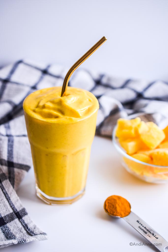 mango turmeric ginger smoothie with black striped towel, bowl of frozen mangoes and spoon of turmeric powder beside it