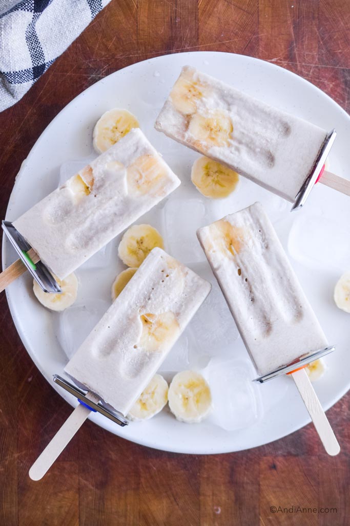 ice pops on white plate with ice cubes and banana slices. Wood cutting board is in background.