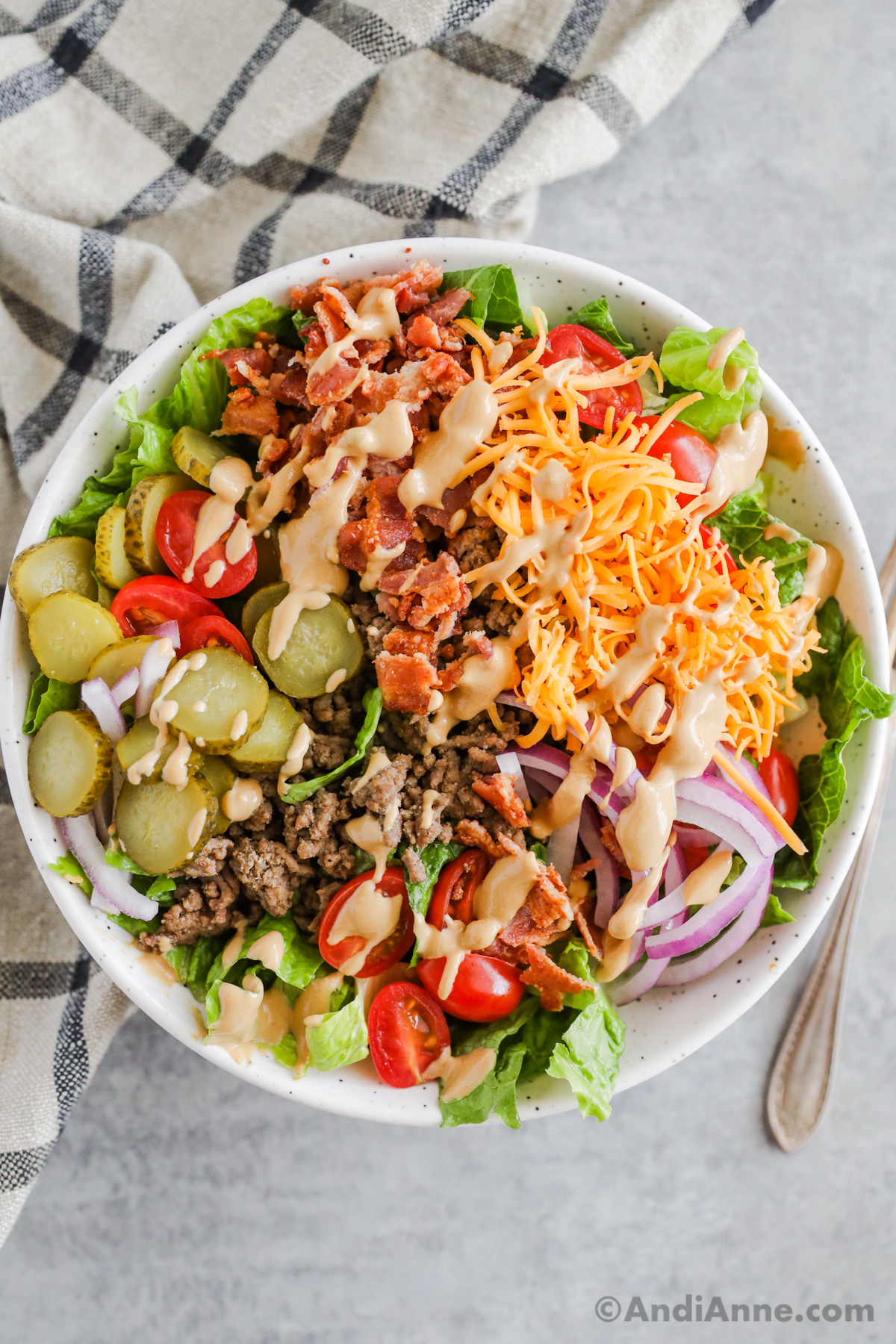 A burger salad bowl recipe with pickles, ground beef, cheese, bacon, onion and drizzled with dressing.