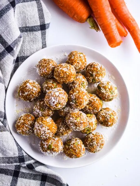 carrot cake bites on a white plate with raw carrots and a kitchen towel surrounding the plate.