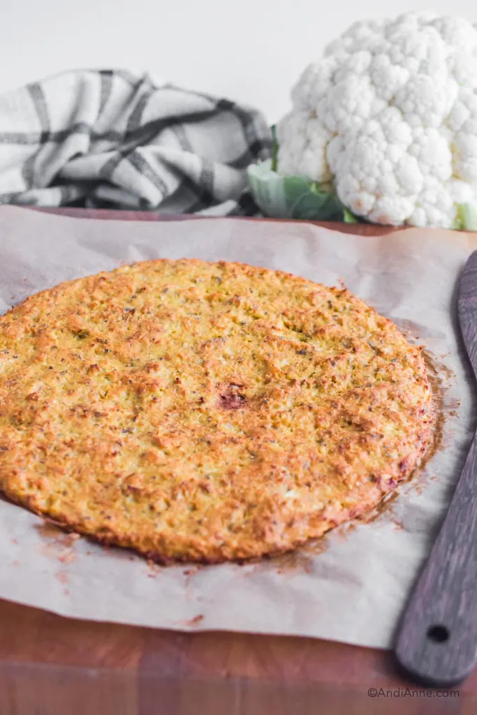 cauliflower pizza crust on parchment paper with wood spatula and cauliflower head in the background