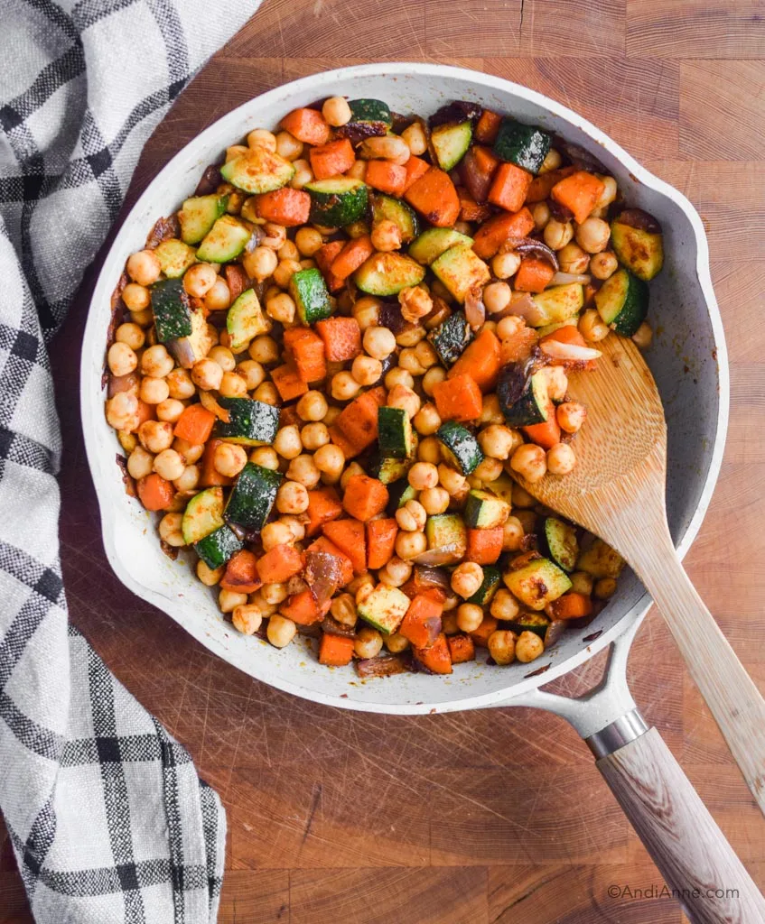 chickpeas, carrots and zucchini in frying pan with wooden spoon and kitchen towel beside it.