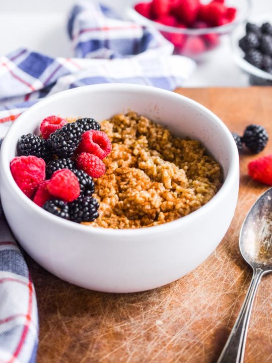 instant pot steel cut oats in a white bowl with fresh berries. Spoon and plaid kitchen towel are beside it.