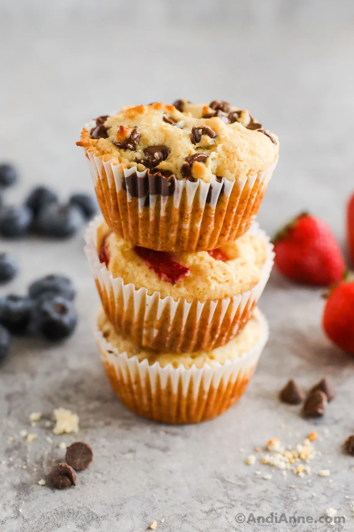 A stack of three pancake muffins with chocolate chips and berries. Behind in background is blueberries, strawberries and some chocolate chips.