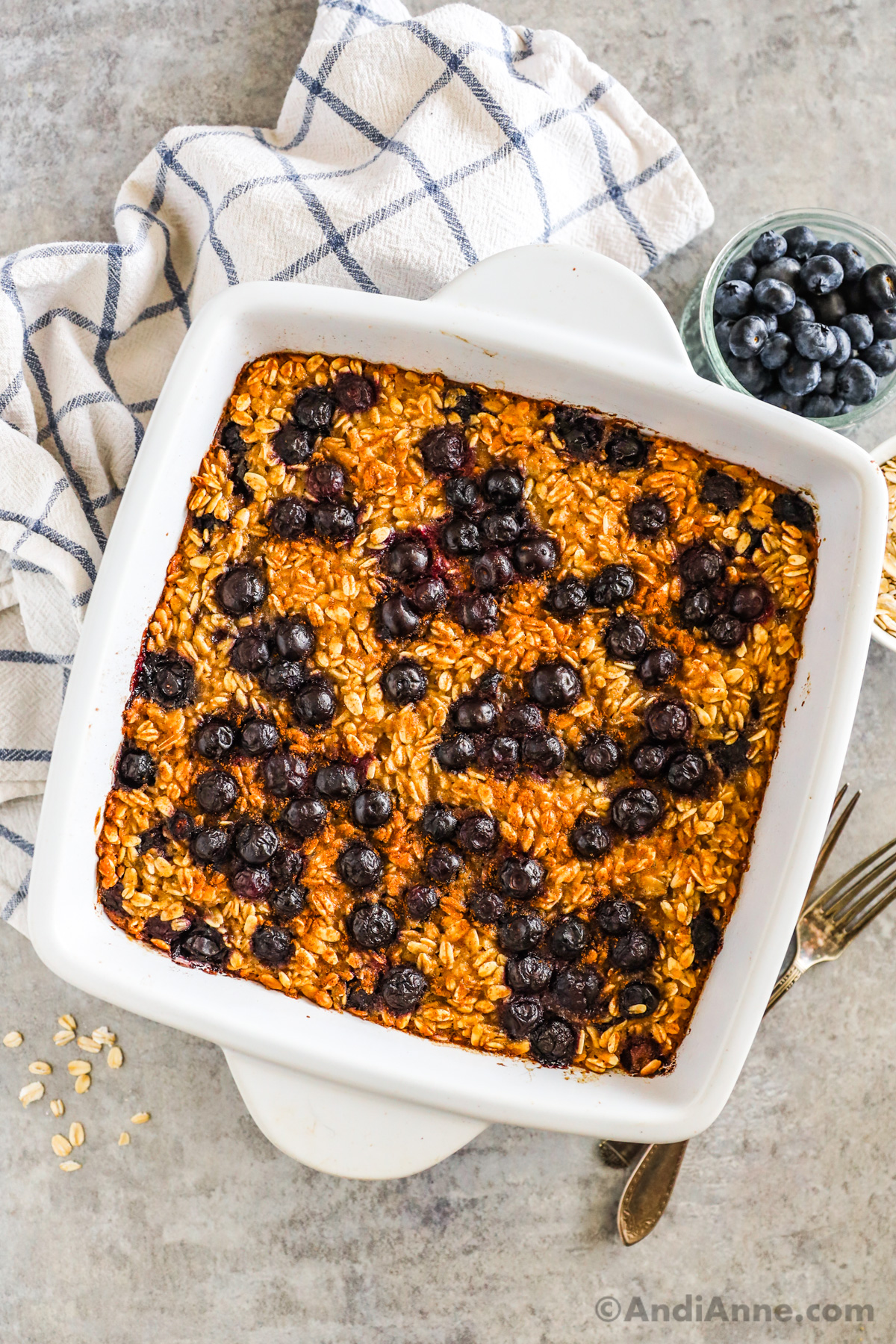A square dish of blueberry baked oatmeal with kitchen towel, forks, and small bowl of fresh blueberries surrounding it.