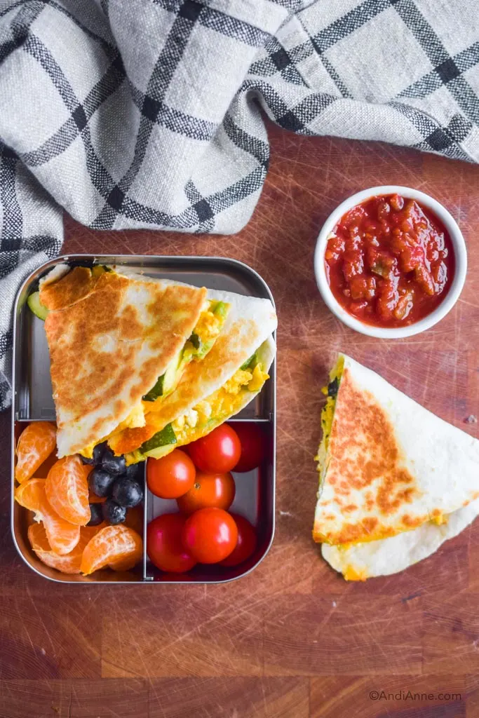 quesadilla in steel container with cherry tomatoes, oranges and grapes. Cup of salsa beside it