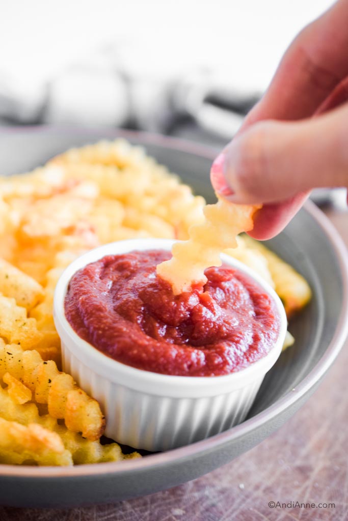 hand dipping a fry into a cup of ketchup with fries behind it