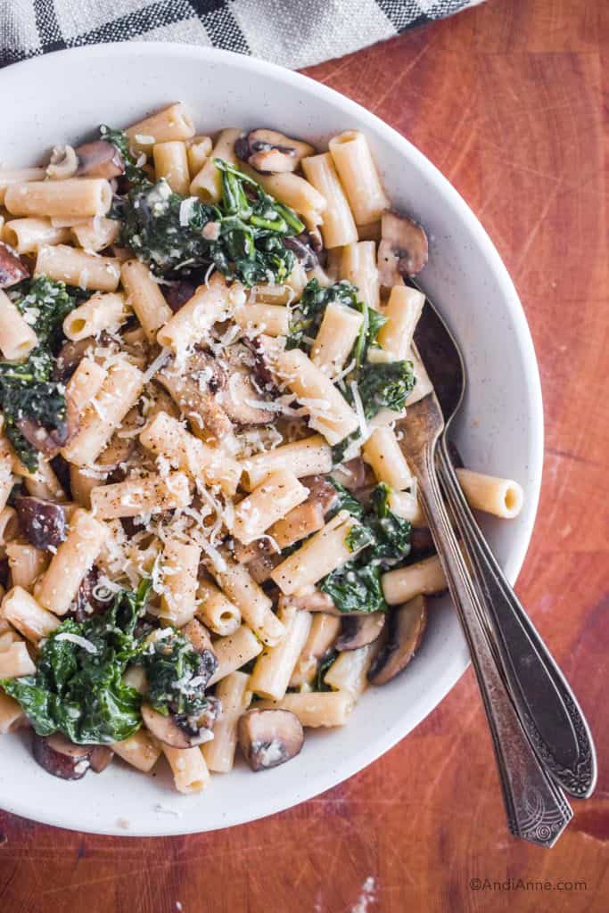 mushroom kale pasta with parmesan cheese in a white bowl. On wood cutting block with kitchen towel in corner.