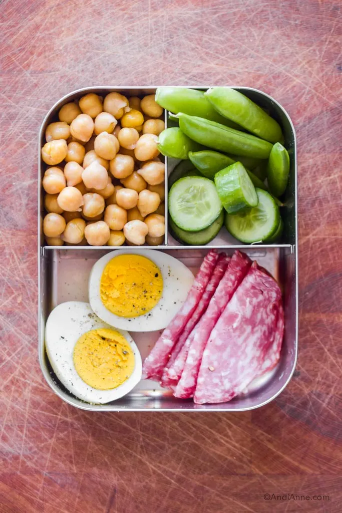 Stainless steel bento box lunch with sliced hard boiled egg, salami slices, snap peas, chickpeas and cucumber slices.