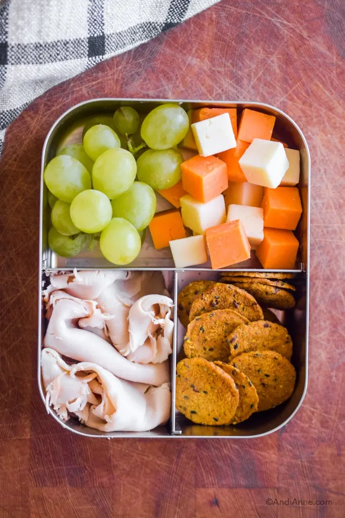 Stainless steel bento box lunch with deli turkey slices, crackers, grapes and cheese cubes.