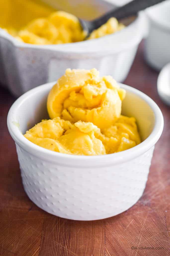 frozen mango ice cream in a white bowl. Dish with rest of mango ice cream and ice cream scoop are in the background.