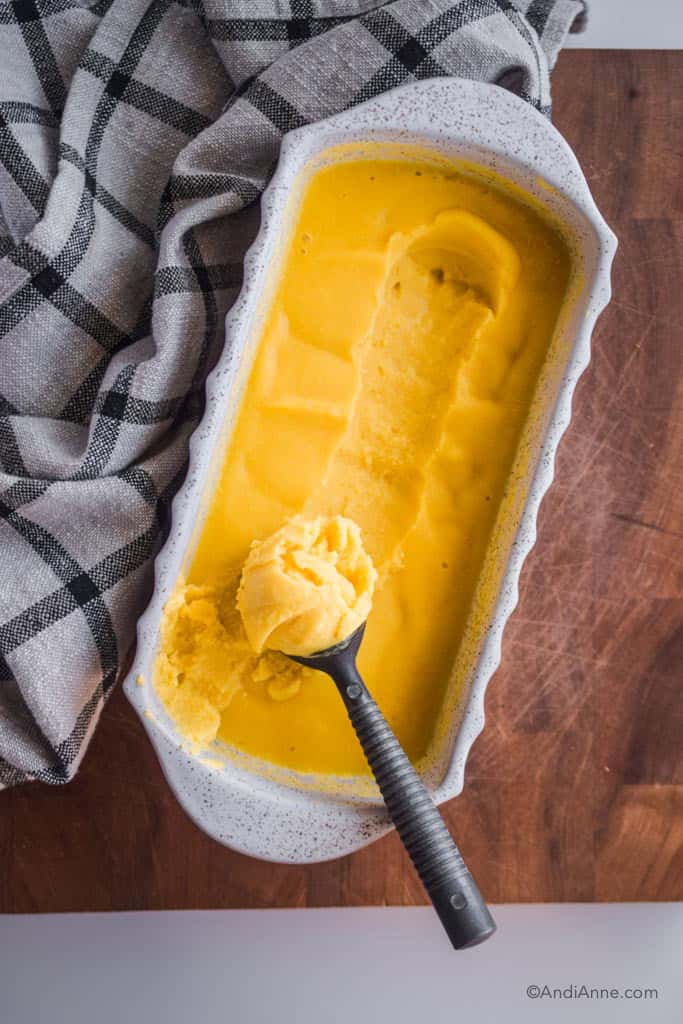 Looking down on a white dish with frozen mango ice cream inside. Once scoop has been rolled out into a silver ice cream spoon.