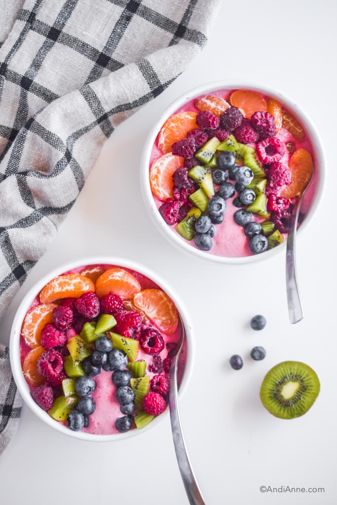 two rainbow smoothie bowls in white bowls with spoons on white table. A slice of kiwi and some blueberries on the white table beside it.