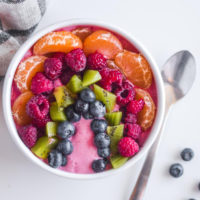 orange slice, raspberries, kiwi and blueberries assembled to create a rainbow on top of a smoothie inside a white bowl with spoon