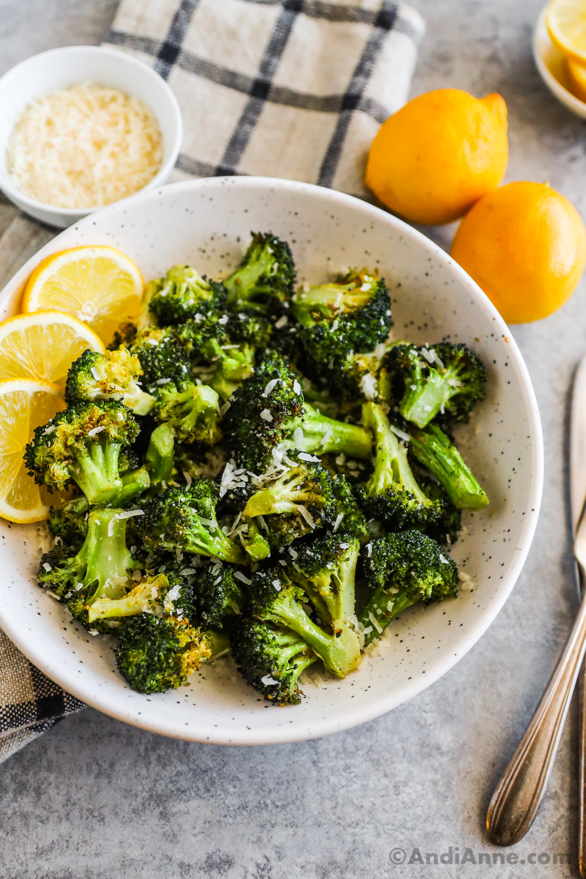 Air fryer broccoli in a bowl with lemon slices and sprinkled with grated parmesan. Fresh lemons and a bowl of grated parmesan surround the bowl.