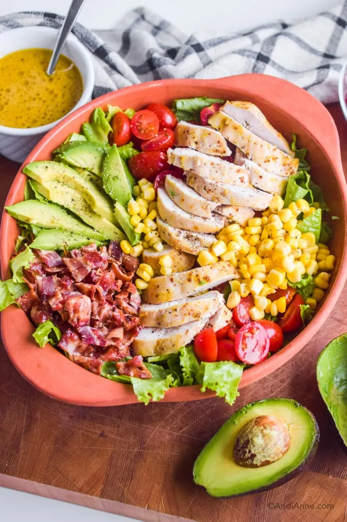 maple dijon chicken avocado salad in bowl. White bowl of dressing with spoon and avocado sliced in half surround it.