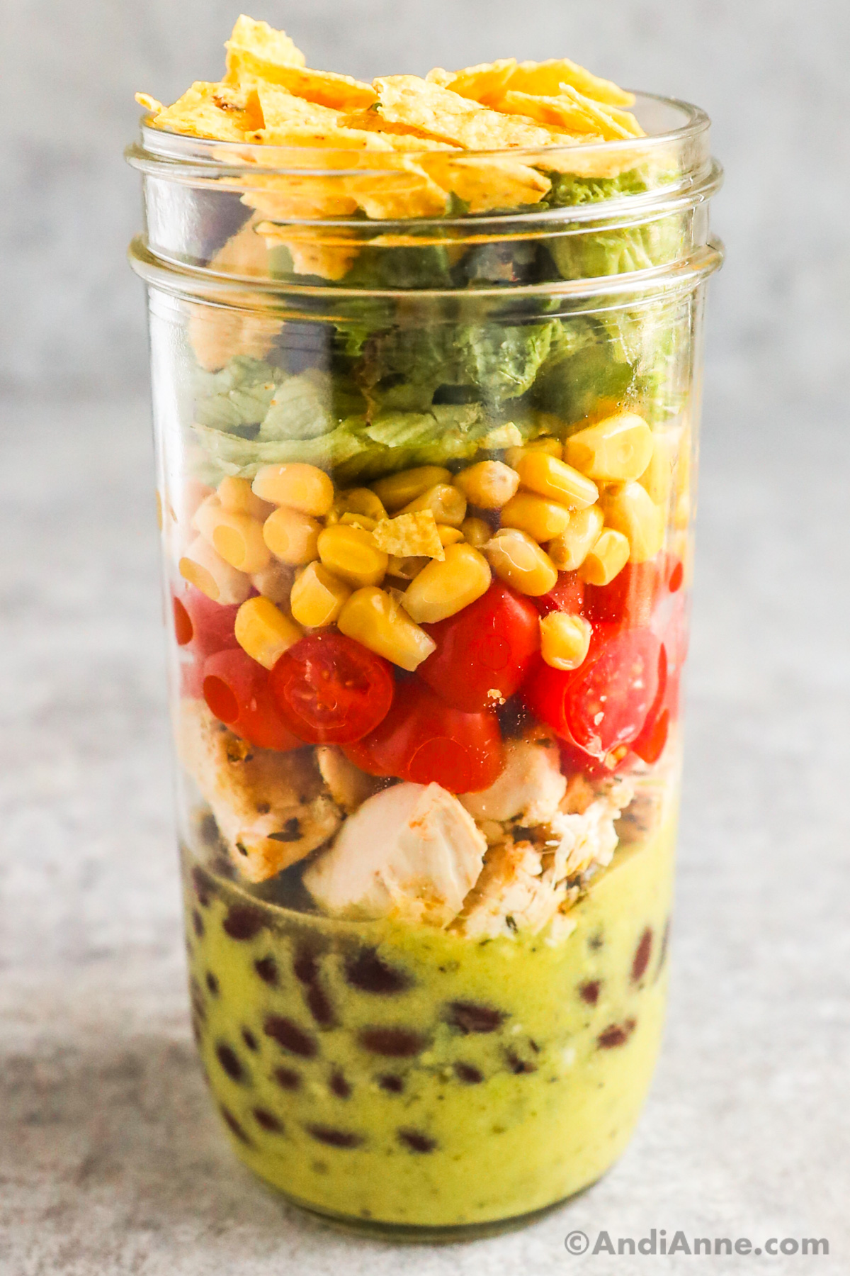 A mason jar salad with layers of dressing, black beans, chicken, tomatoes, corn, mixed greens and crushed tortilla chips.