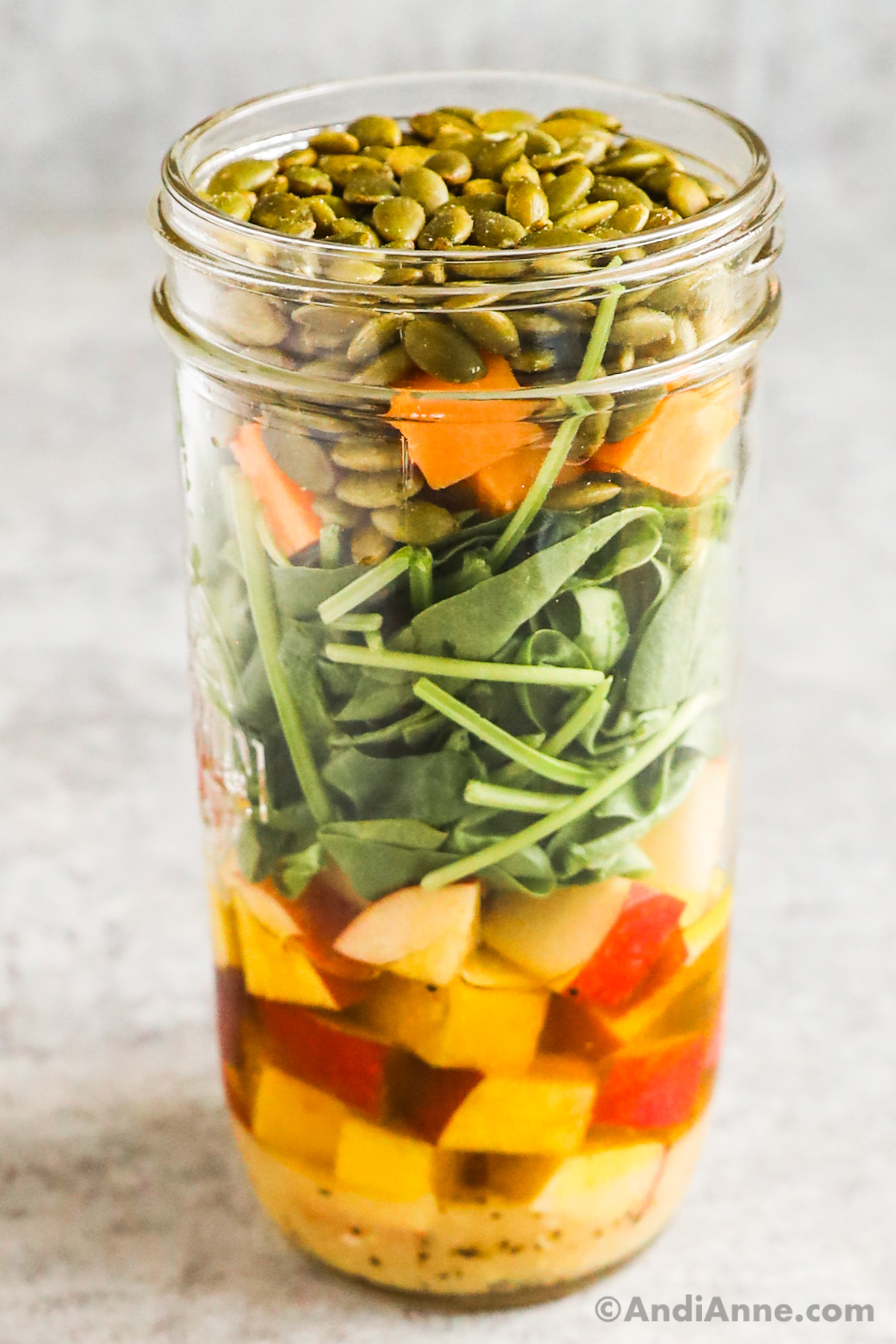 A mason jar salad with layers of dressing, chopped apple, spinach, cheese and pumpkin seeds.