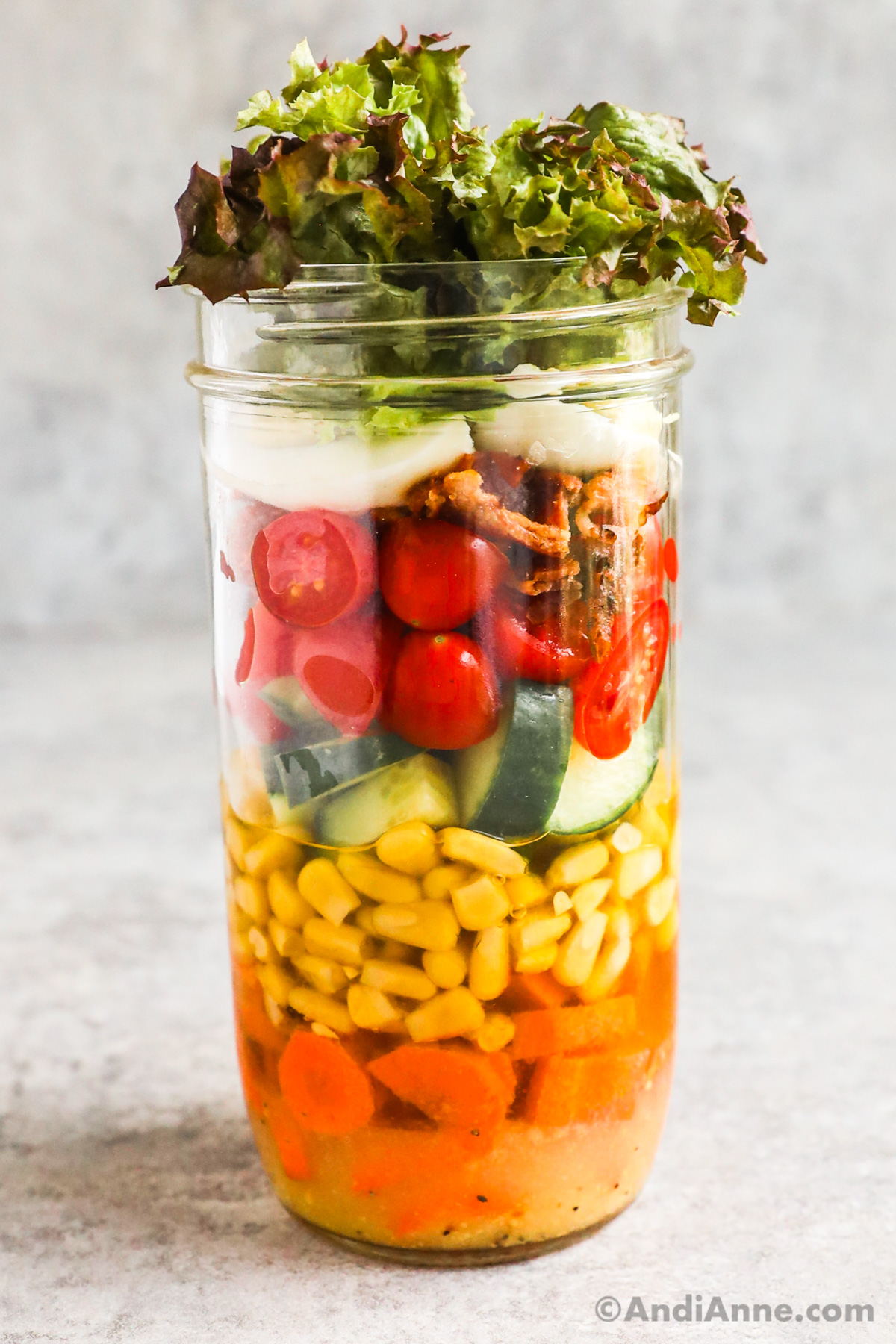 A cobb mason jar salad with layers of dressing, carrots, corn, cucumber, tomatoes, bacon, egg and mixed greens.