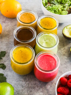 Six different homemade salad dressings in small jars surrounded by colorful fresh fruits and vegetables