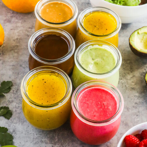Six different homemade salad dressings in small jars surrounded by colorful fresh fruits and vegetables
