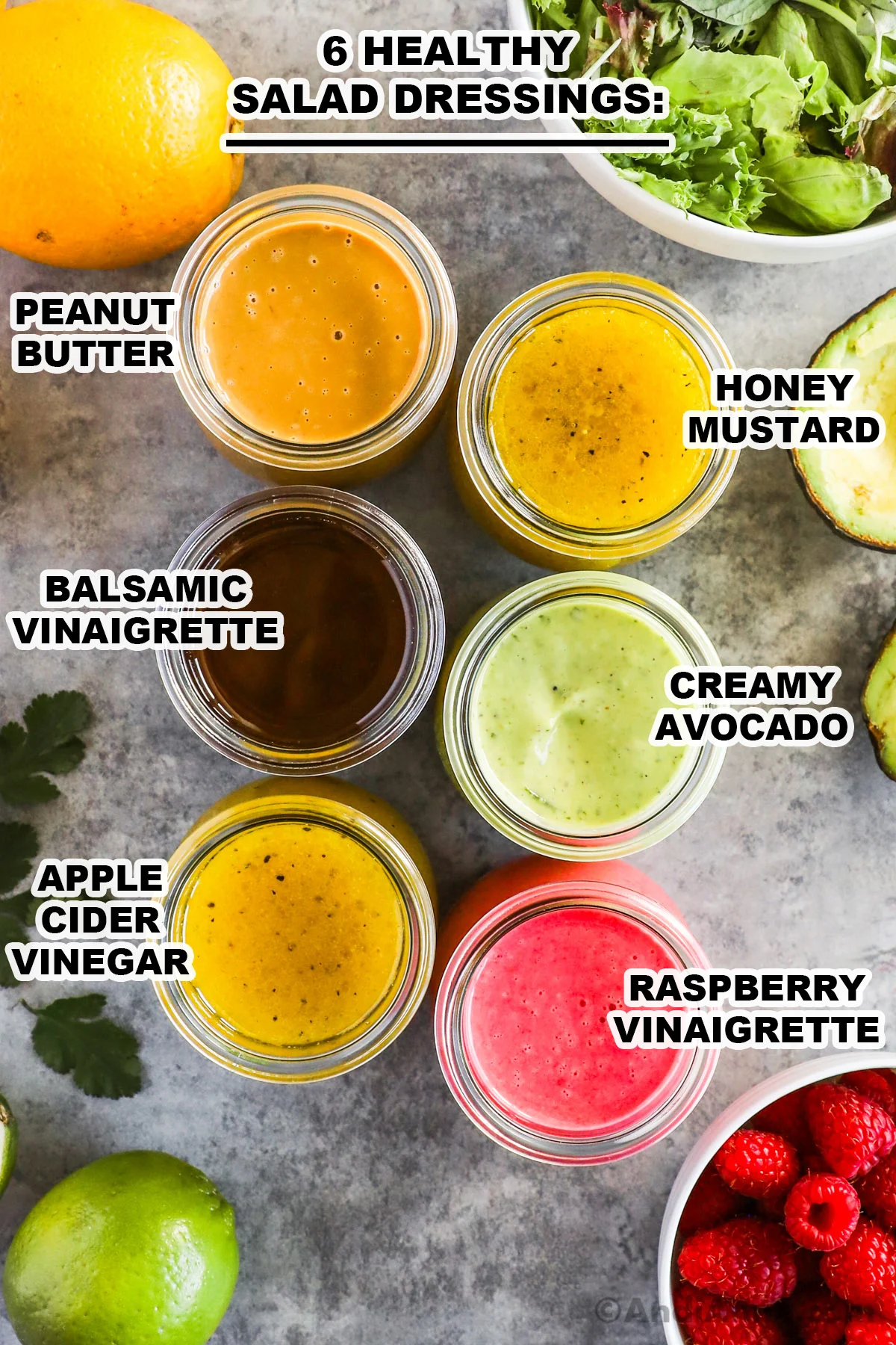 Six colorful jars of homemade salad dressing surrounded with vegetables and fruit.
