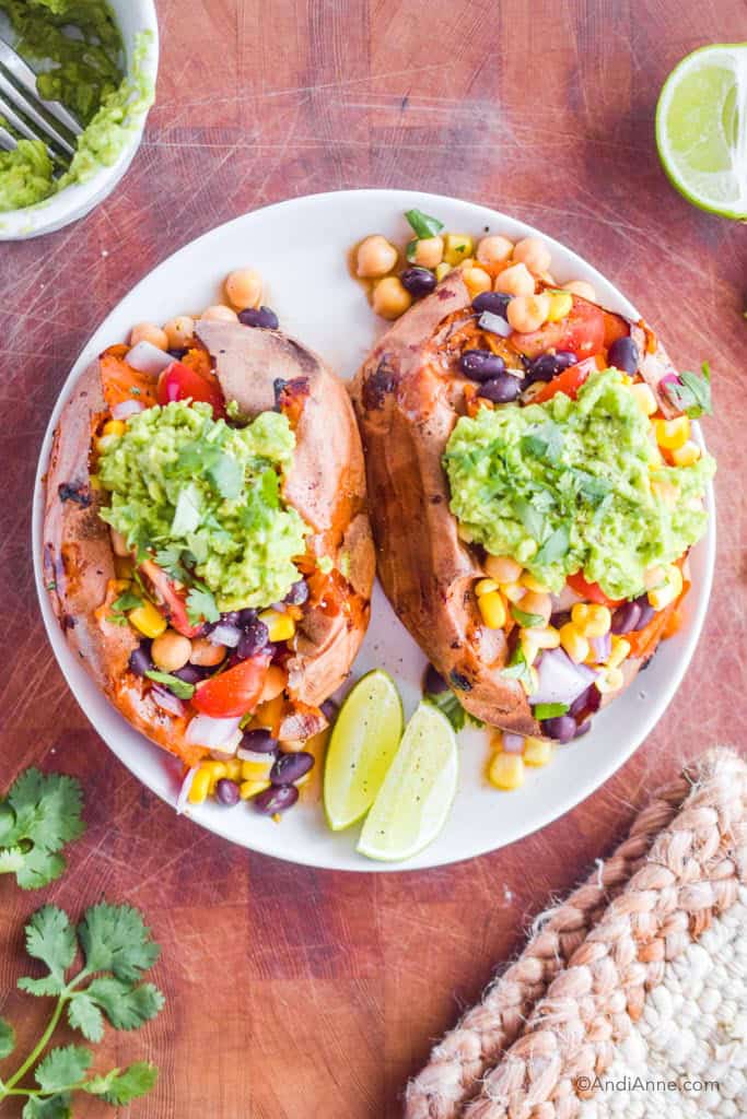 taco stuffed sweet potaoes on a white plate with sliced lime. Cilantro, mashed avocado in a bowl, limes and a napkin surround the plate.