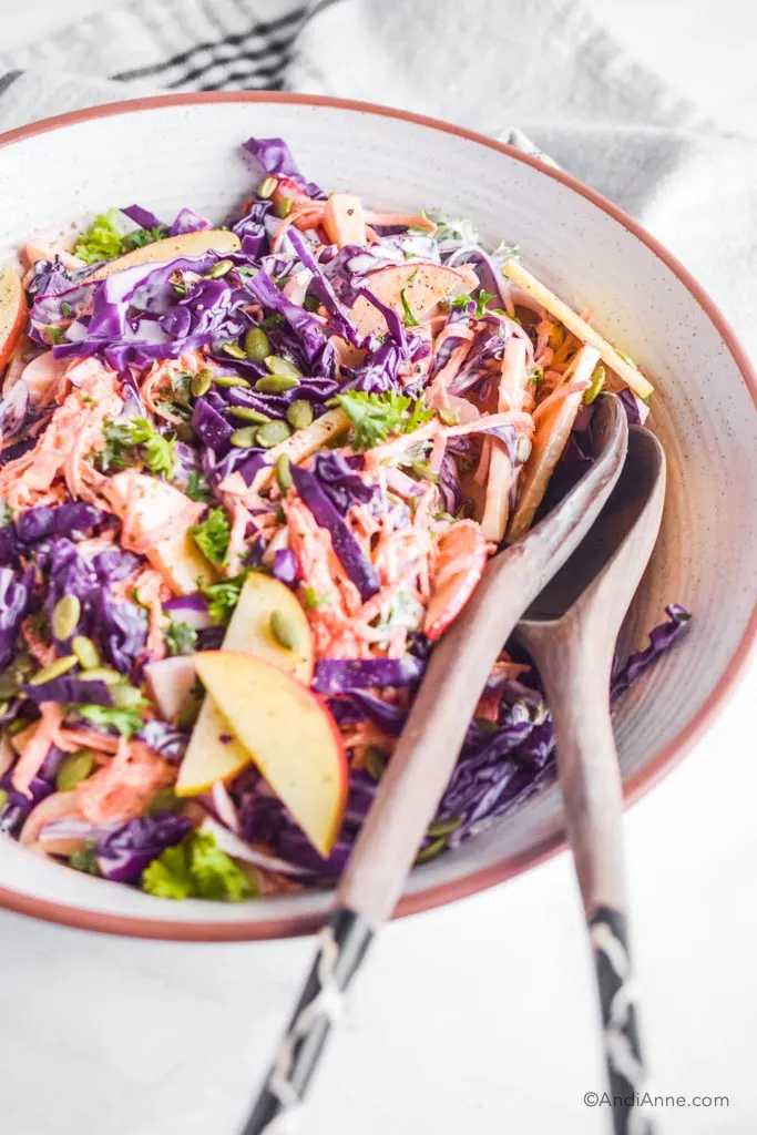 purple cabbage, carrot, apple coleslaw in a white bowl with wood salad spoons.