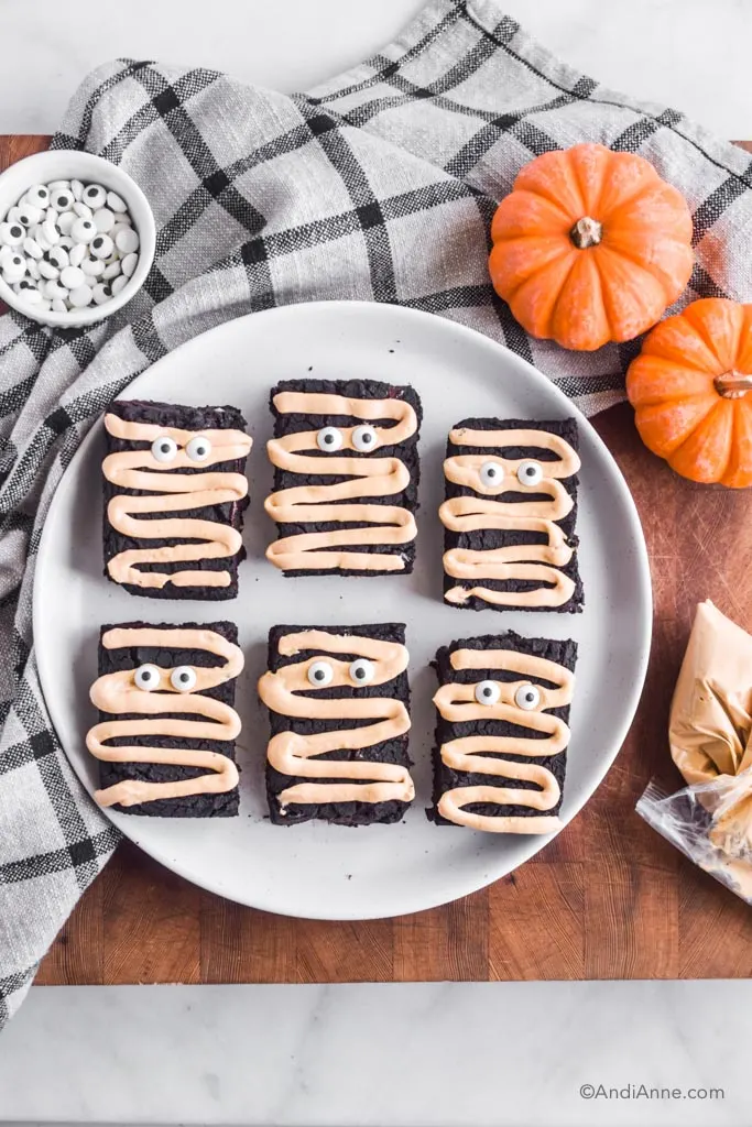 Looking down at the white plate of halloween brownies with two small pumpkins, a piping bag of light brown cream cheese icing, and a bowl of candy eyeballs, and a plaid kitchen towel surrounding the plate.