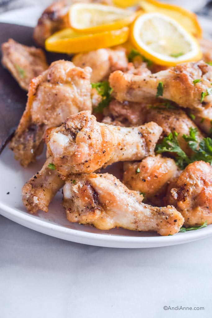 Close up of lemon garlic chicken wings on a white plate with parsley and lemon slices blurred in background