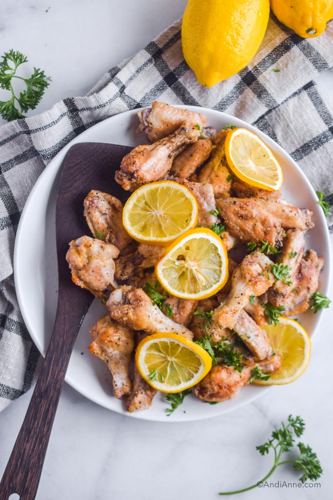 lemon garlic chicken wings on a white plate with wooden spoon. Sliced lemon and bits of parsley garnish the chicken.