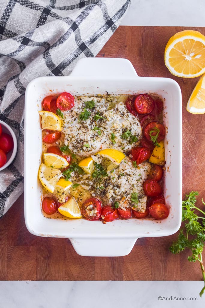 lemon herb baked cod with tomatoes and lemon wedges in a square baking dish. Dish is on a wood cutting board with kitchen towel, sliced lemon and parsley surrounding it. 