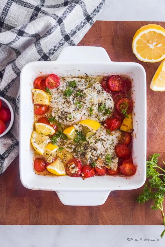 lemon herb baked cod with tomatoes and lemon wedges in a square baking dish. Dish is on a wood cutting board with kitchen towel, sliced lemon and parsley surrounding it.