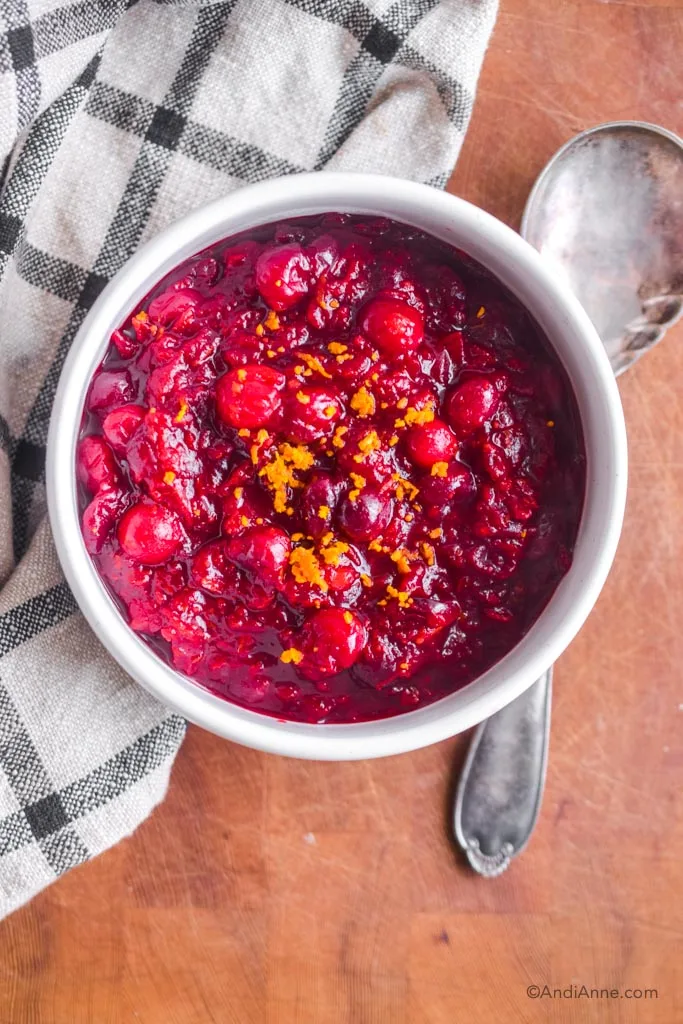 cranberry sauce with orange zest in a white bowl with silver spoon and kitchen towel beside the bowl