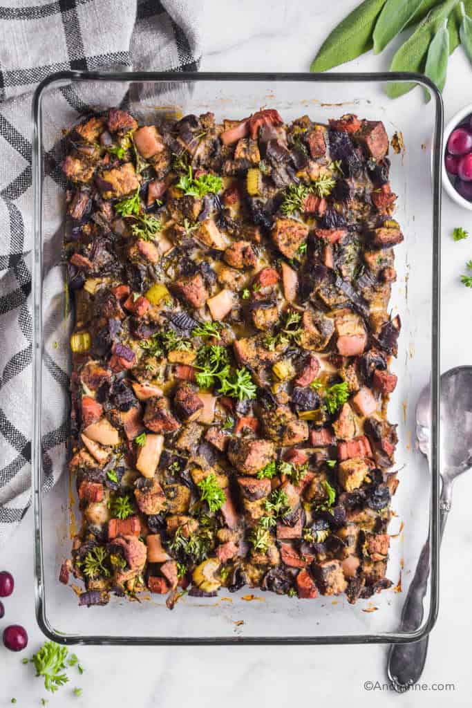 keto stuffing in a glass casserole dish. A kitchen towel, silver spoon, small bowl of cranberries, and fresh sage surround the dish.