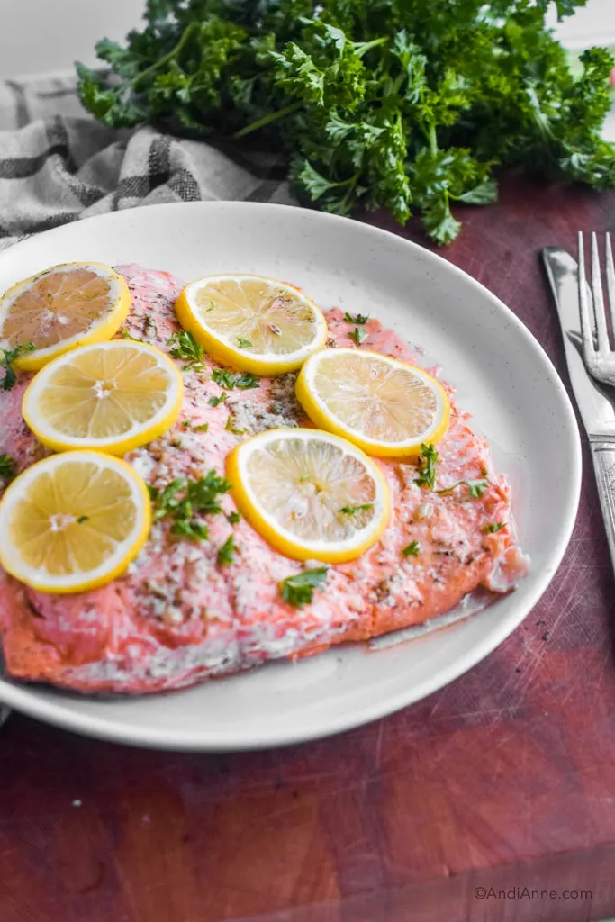 Cooked steelhead trout on a white plate with lemon slices on top. Fork and knife and bunch of parsley surround the plate.