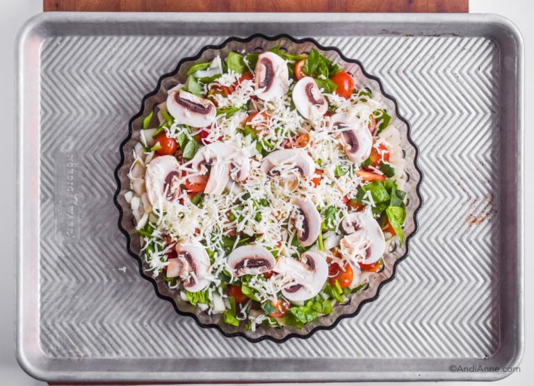 Raw sliced mushrooms, tomatoes and spinach in a baking dish with shredded cheese on top.